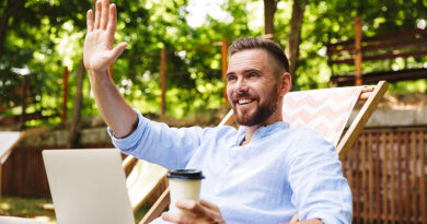 Photo of smiling emotional young bearded man outdoors using laptop computer looking aside waving holding coffee.