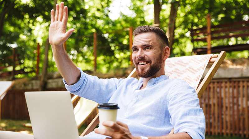 Photo of smiling emotional young bearded man outdoors using laptop computer looking aside waving holding coffee.