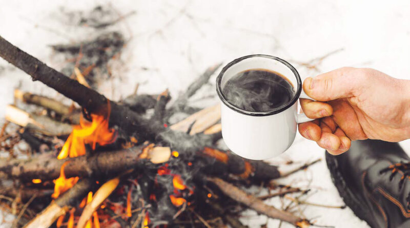 Male hands hold a mug of coffee near a burning campfire. Concept hike, walk, trip in winter